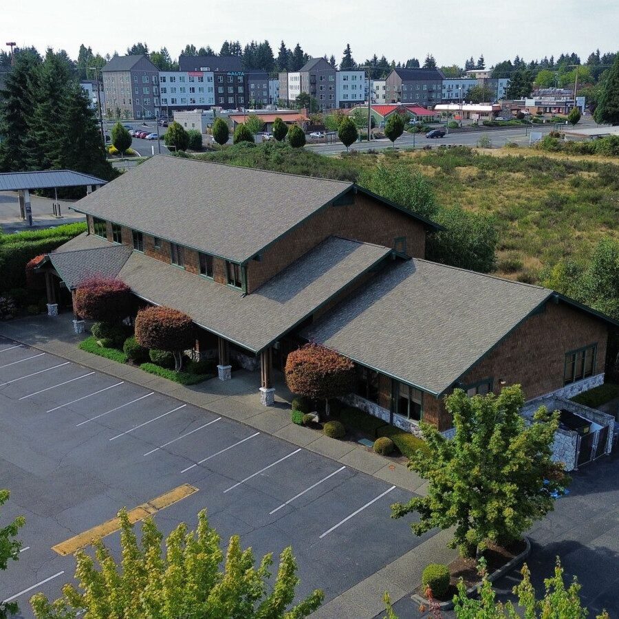A two-story building with a sloped roof sits next to an empty parking lot, surrounded by trees and bushes, with commercial buildings and South Sound commercial property management services nearby.