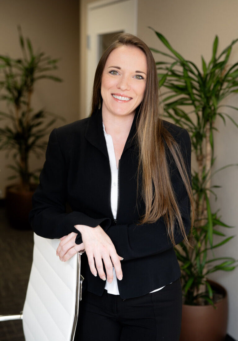 A woman with long brown hair, dressed in a black blazer and white shirt, stands in an office next to a white chair, with potted plants in the background.