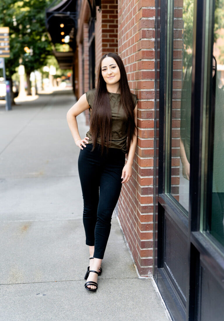 A woman with long brown hair, wearing a green top, black pants, and sandals, stands against a brick wall on a city sidewalk, looking at the camera.