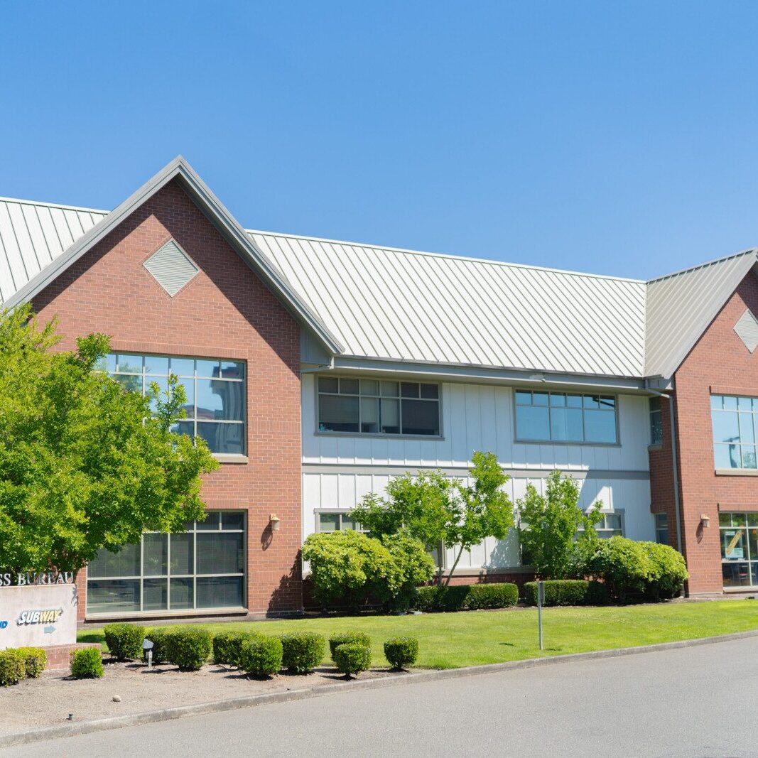 A two-story brick and white commercial building with large windows, surrounded by trimmed bushes, trees, and a lawn under a clear blue sky. A sign reads “Bellinger Business Center,” managed by a leading South Sound property management company.
