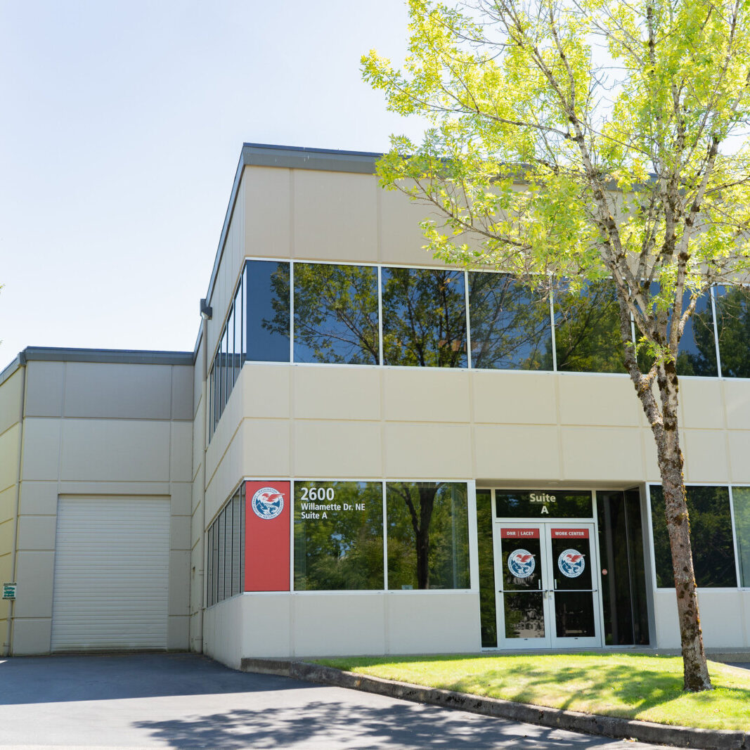A two-story commercial building with large windows, a red business sign, and a tree in front, photographed on a clear, sunny day.