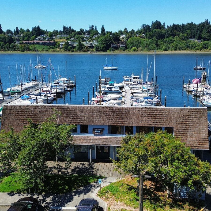 A marina with numerous boats docked in the water, a wooden building in front, trees, parked cars, and houses across the river under a clear blue sky—perfect for those seeking guidance from a South Sound property management company.