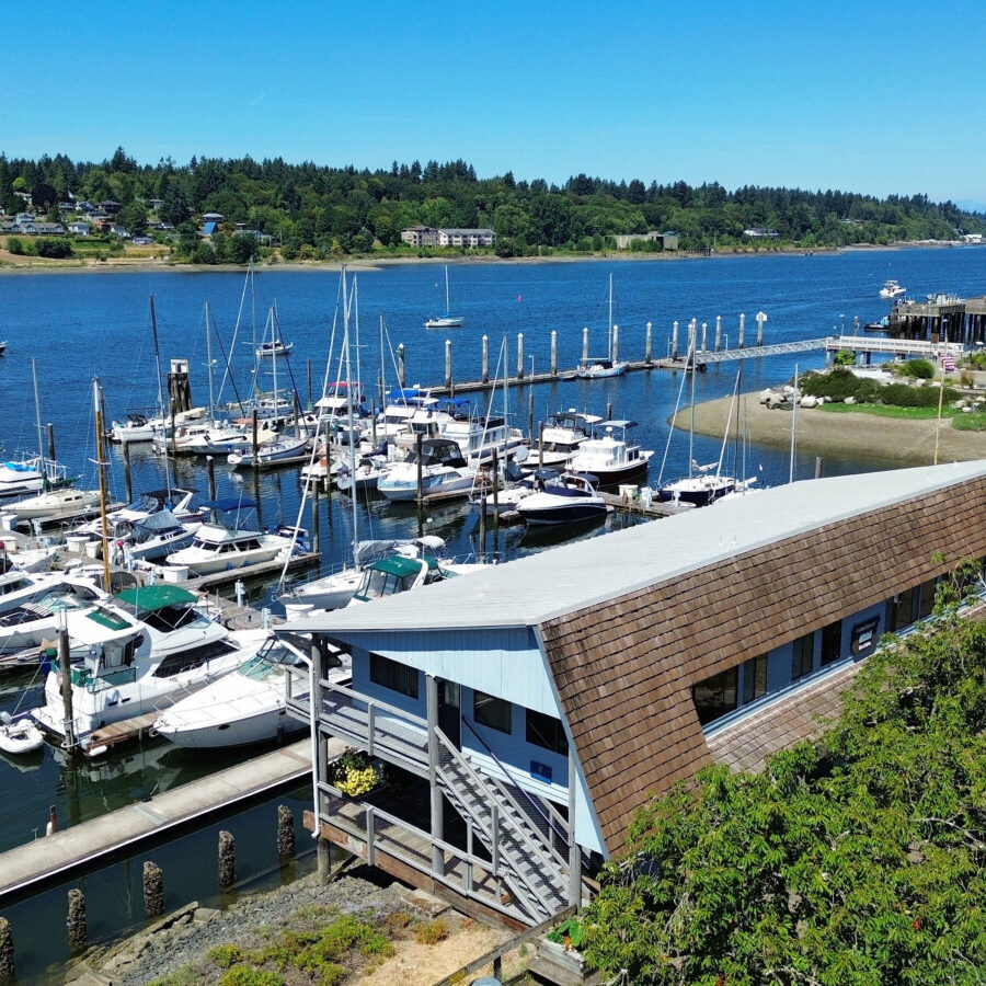A marina with several docked boats and a long building in the foreground, bordered by trees and water, under a clear blue sky.