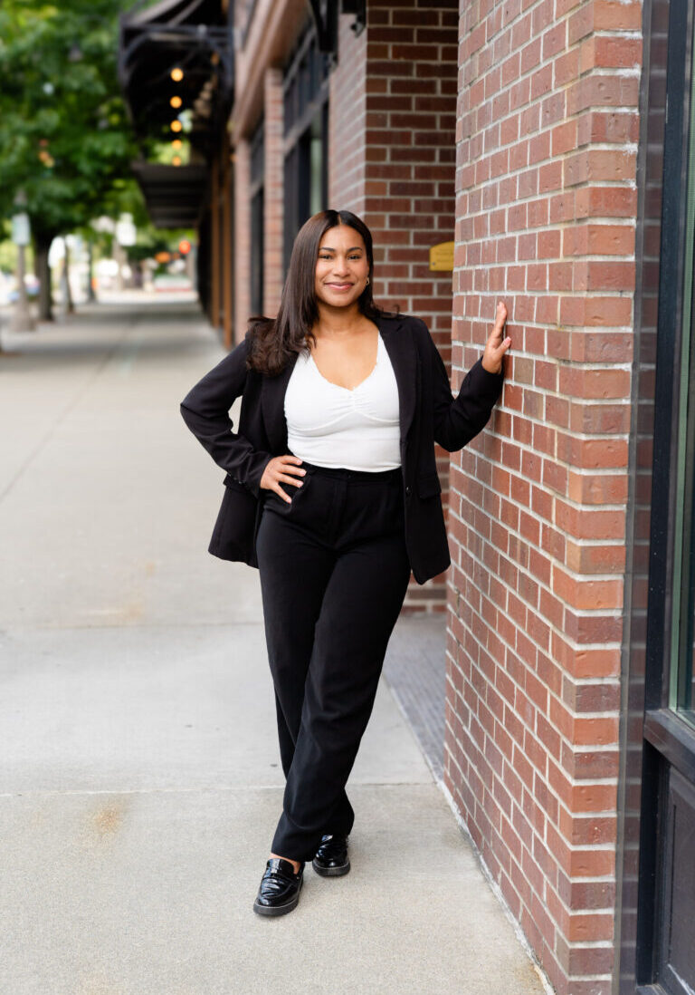 A woman wearing a black suit and white top stands confidently against a brick wall on a city sidewalk, with one hand resting on the wall and a slight smile.