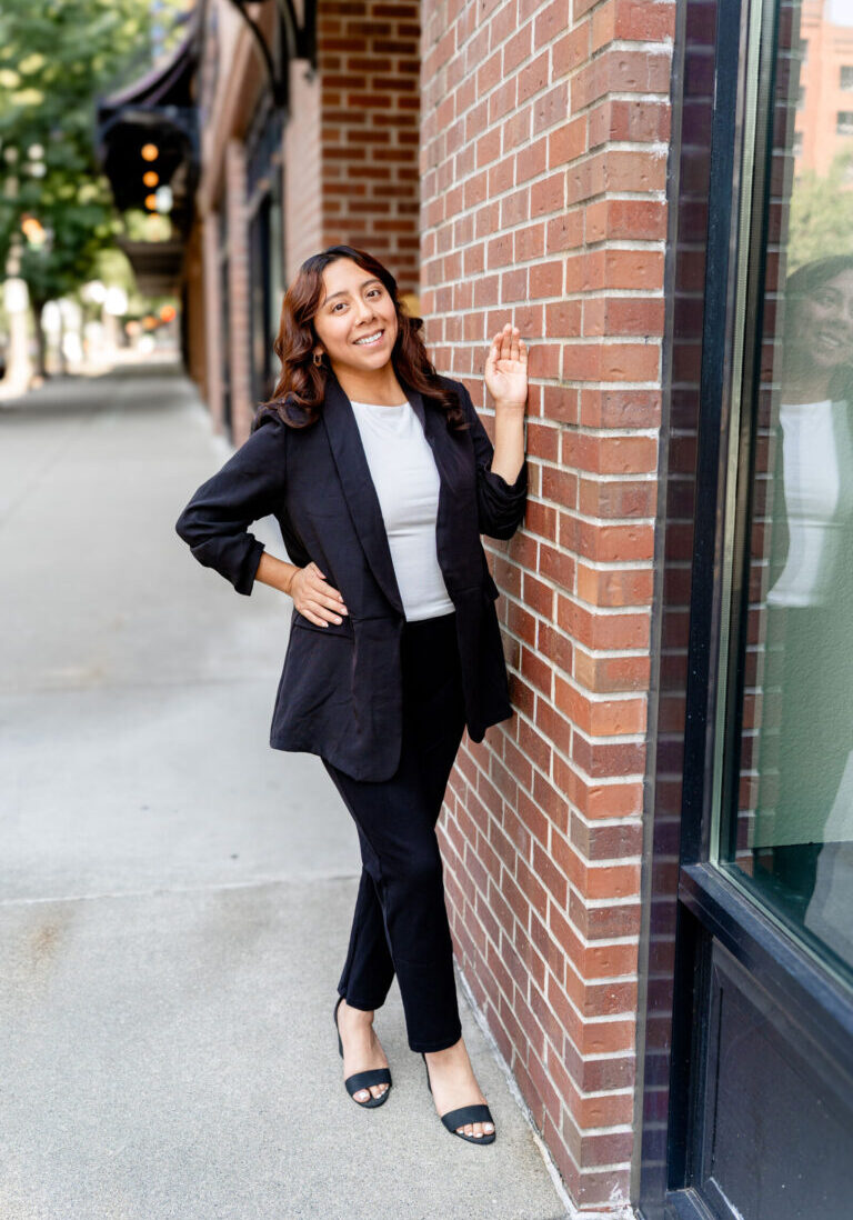 A woman in a black suit and white top stands against a brick wall on a city sidewalk, smiling with one hand resting on the wall.
