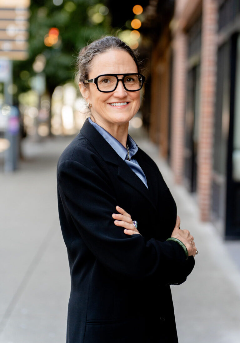 A woman wearing glasses and a black blazer stands outdoors on a city sidewalk, arms crossed, smiling at the camera.