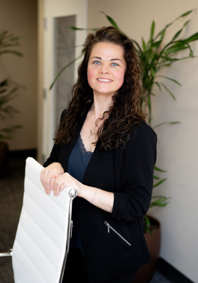 A woman with curly brown hair, wearing a black blazer and navy top, stands in an office holding the back of a white chair. There are green plants and a glass door in the background.
