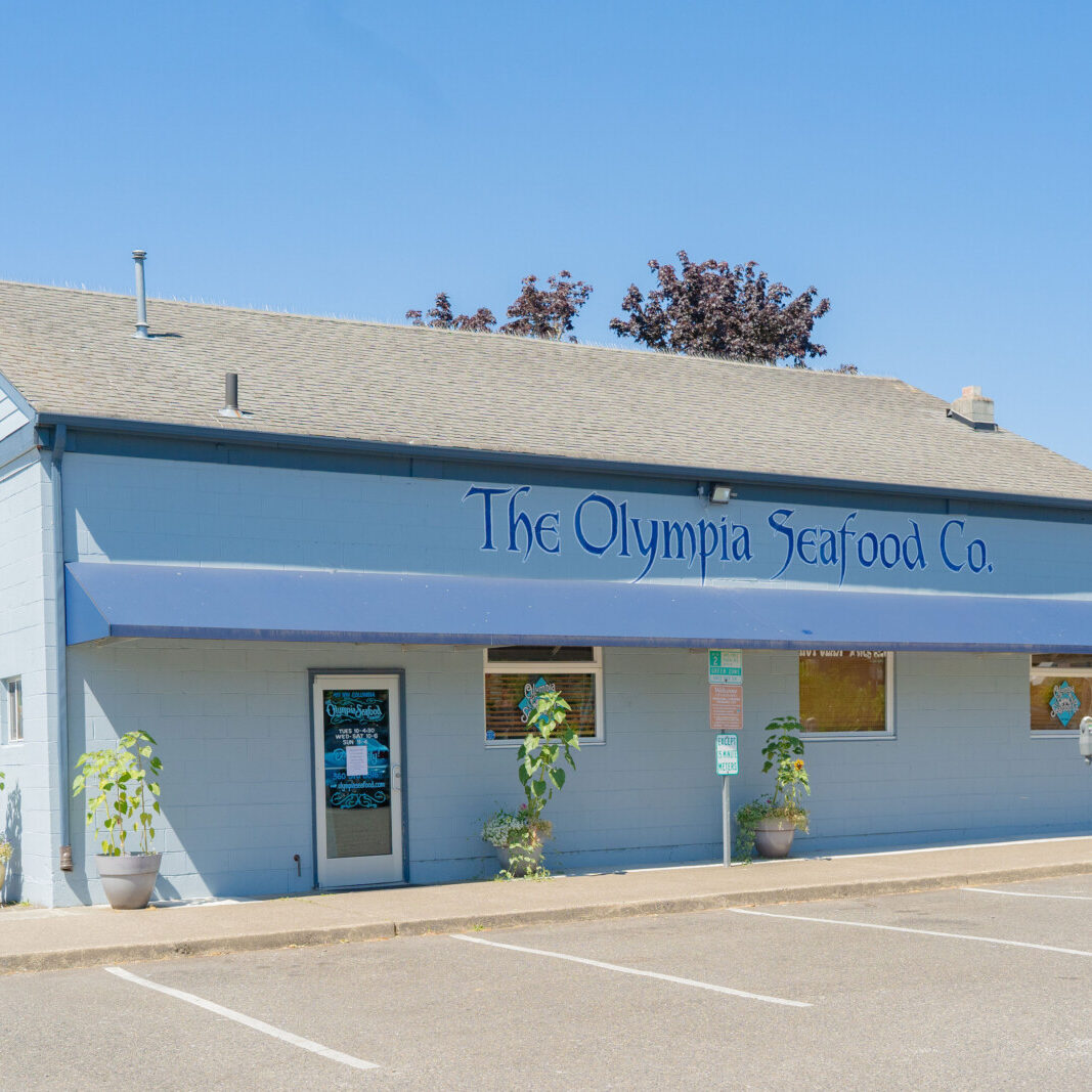 A light blue building with a sign reading "The Olympia Seafood Co." and a blue awning, with potted plants in front and a parking lot in the foreground.