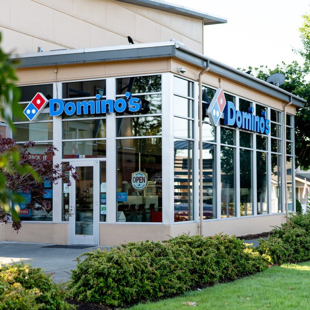 A Domino’s Pizza storefront with large glass windows, an open sign on the door, and lush greenery—an example of South Sound commercial property management services on a sunny day.