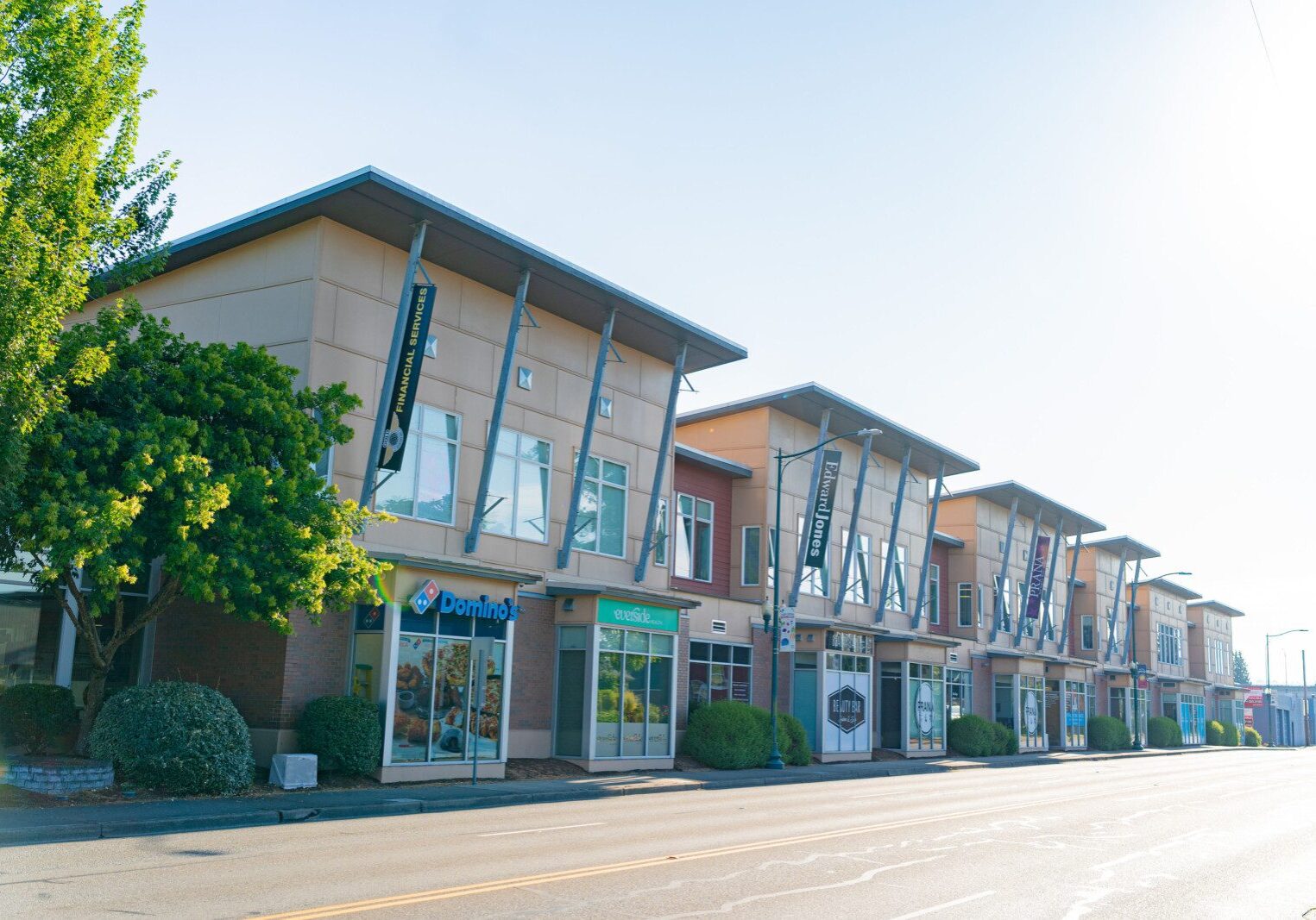 A row of modern commercial buildings with signs, including Domino’s, along an empty street on a sunny day.