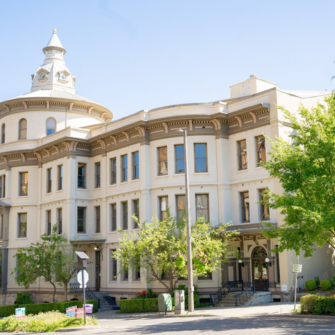 A historic, three-story beige building with a rounded corner tower and large windows offers South Sound commercial tenant services, all nestled among surrounding trees on a sunny day.