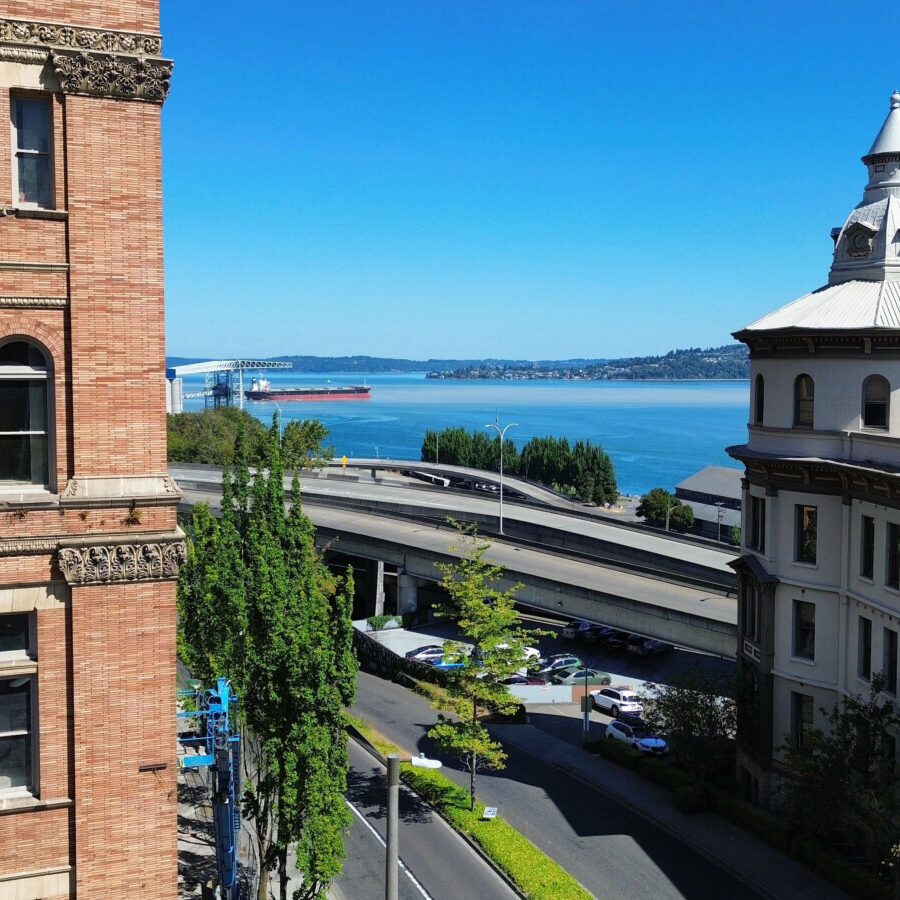 A view of two historic buildings with a road and trees between them, overlooking a body of water where a cargo ship sails—a prime example of South Sound commercial tenant services thriving under a clear blue sky.