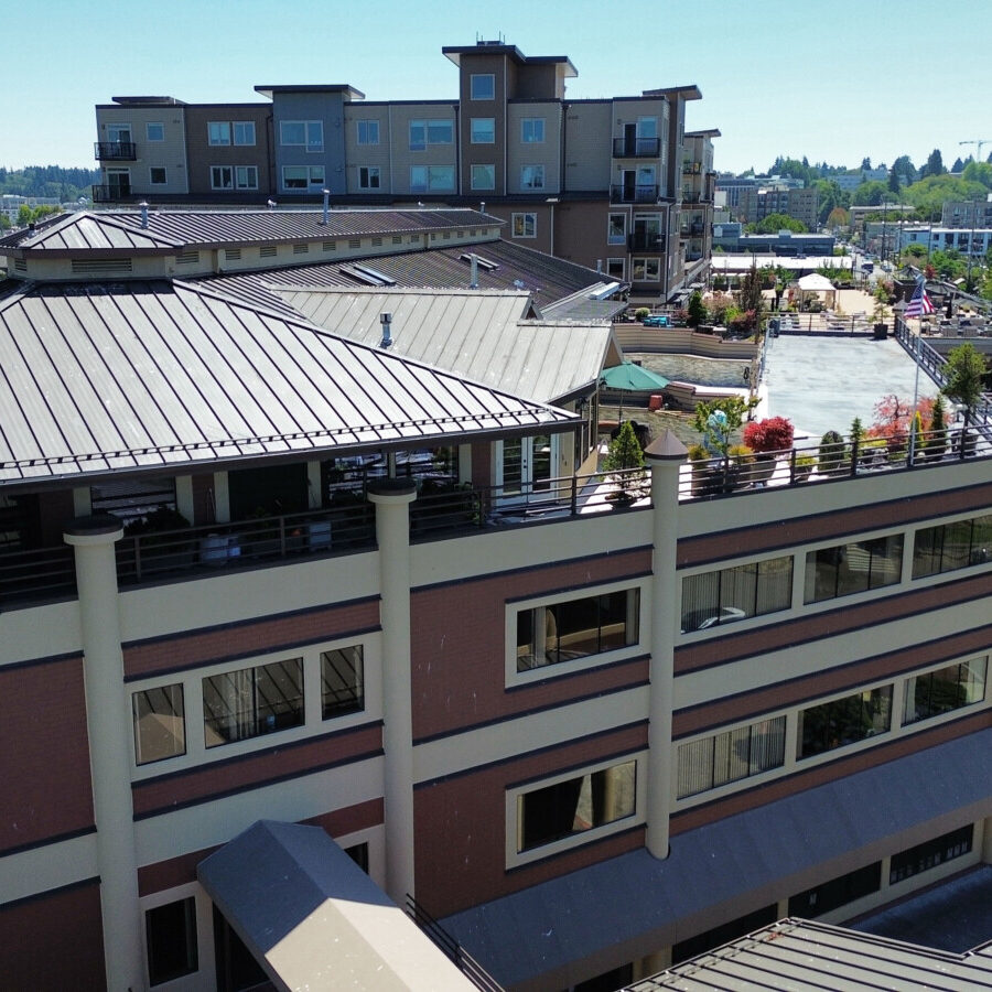 A multi-story modern building with rooftop dining, surrounded by trees and parking, offers South Sound commercial tenant services, with a capitol dome visible in the background under a clear sky.