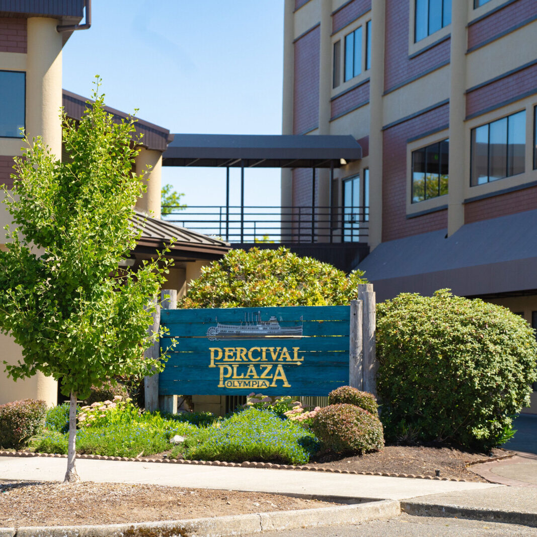 A blue wooden sign for Percival Plaza stands in front of a landscaped area and a multi-story office building at 626, highlighting South Sound commercial tenant services, with part of a blue car visible on the right.