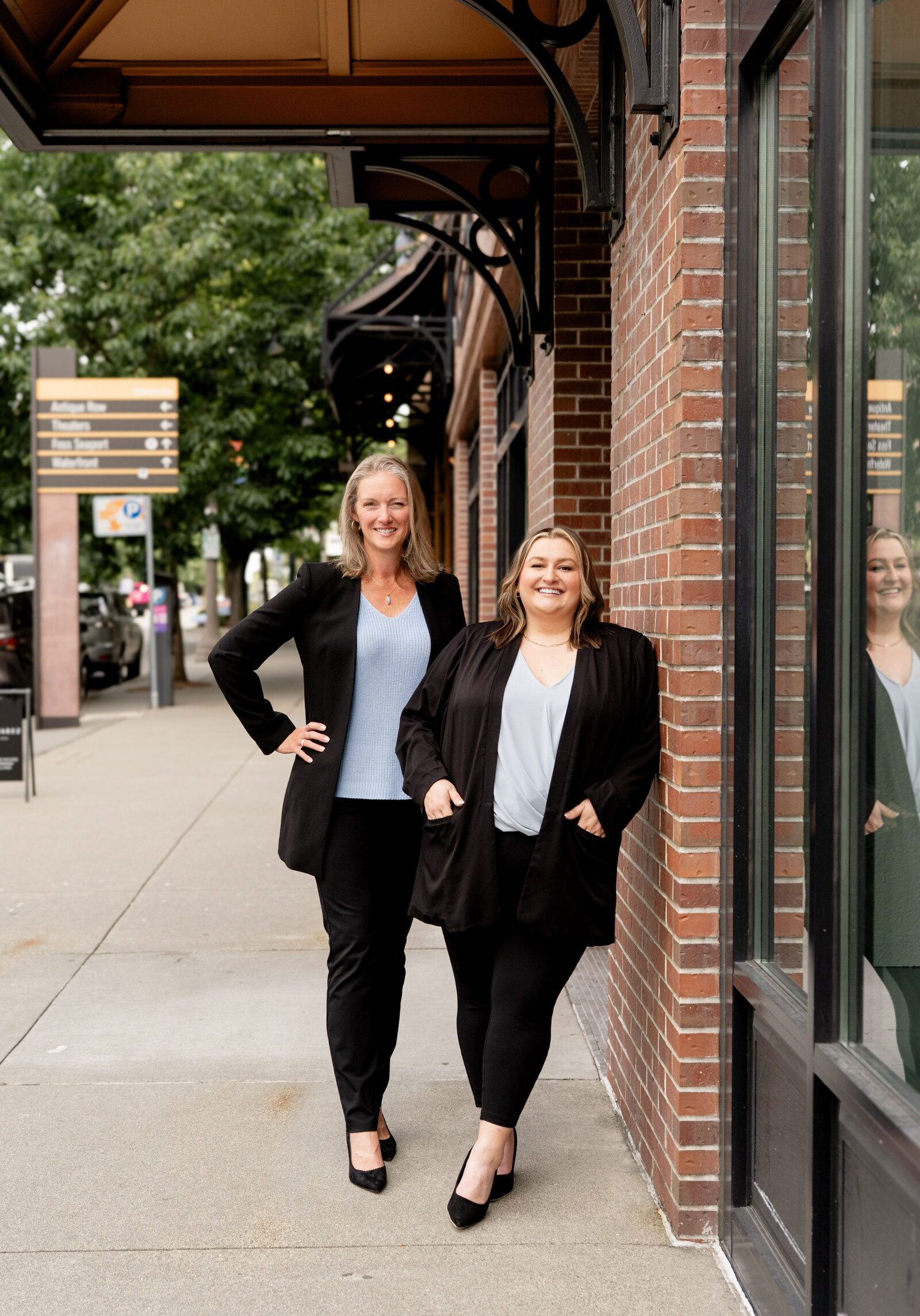 Two women in business attire stand and smile outside on a sidewalk next to a brick building and large window, representing a South Sound property management company, with trees and signs visible in the background.