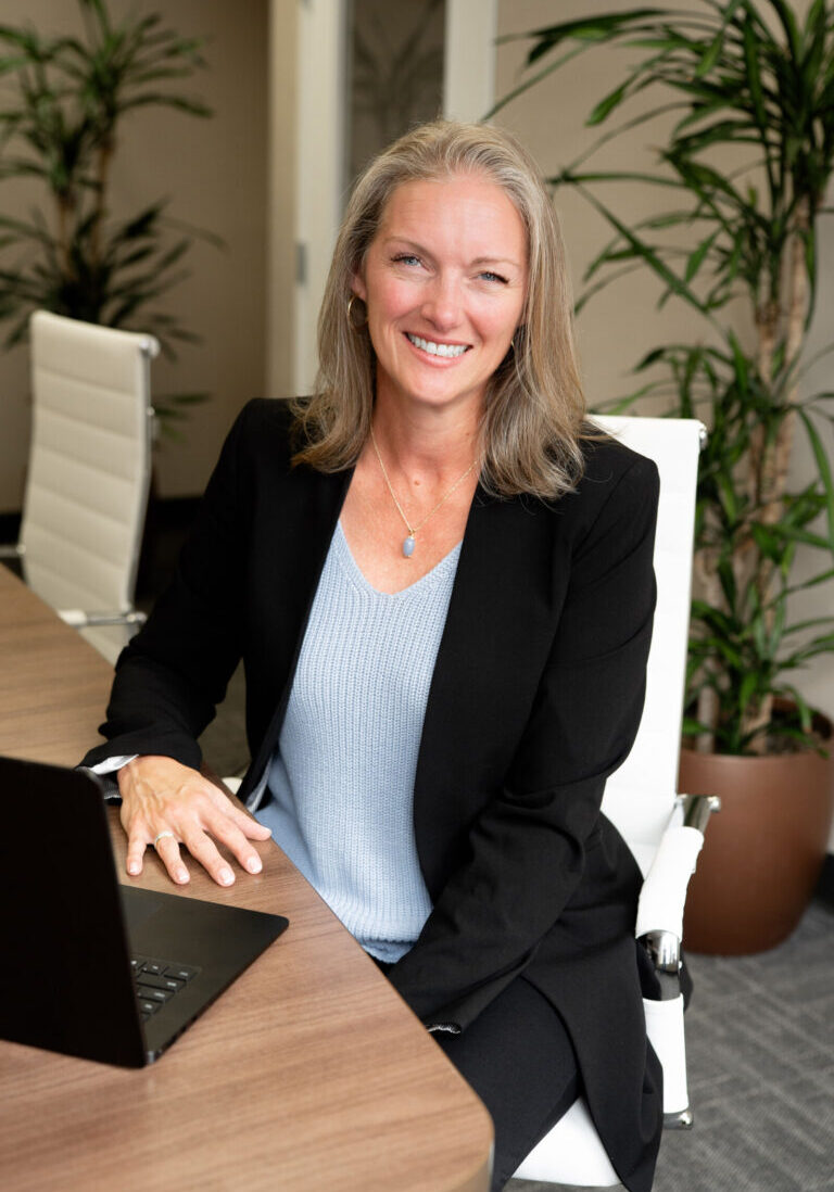 A woman with shoulder-length gray hair in a black blazer and light blue top sits at a desk with a laptop, smiling at the camera in a modern office.