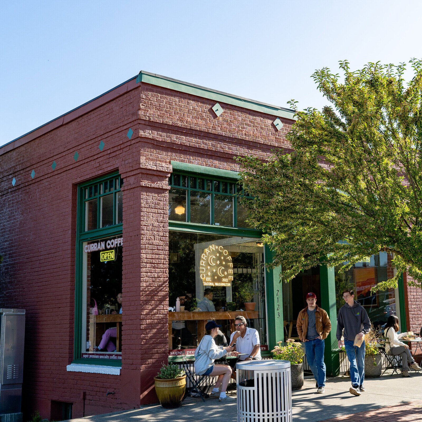 A brick coffee shop with green trim on a sunny day, people sitting and standing outside, and a tree partially shading the sidewalk.