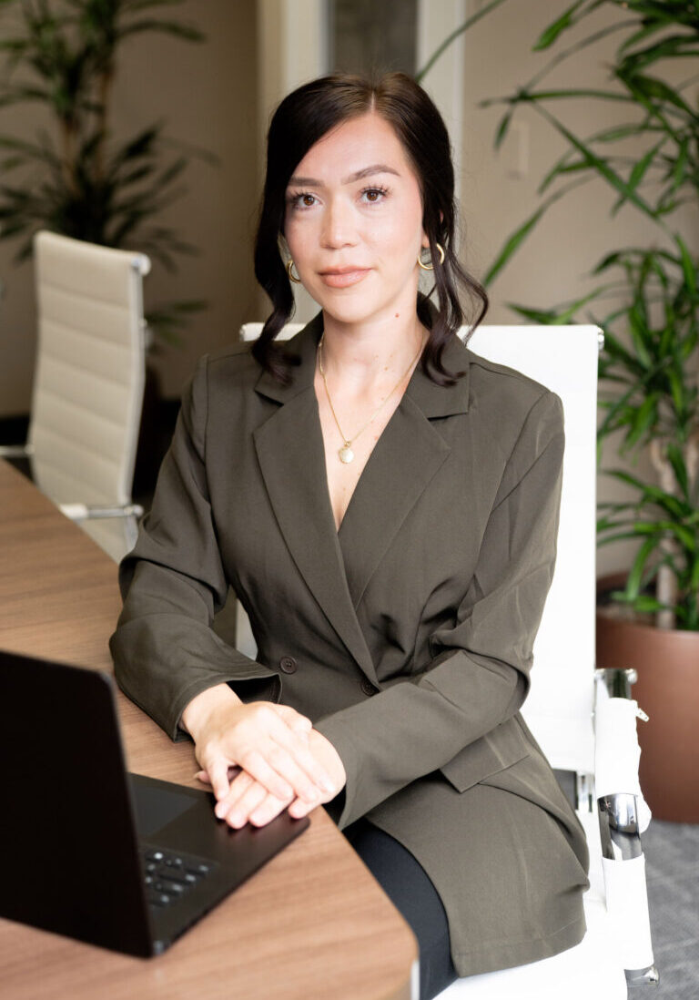 Woman with dark hair sits at a conference table in an office, wearing a dark blazer and gold jewelry, with a laptop in front of her and green plants in the background.